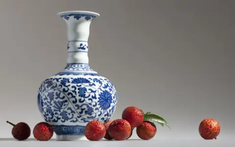 Un vase Satsuma bleu et blanc orné de motifs floraux, entouré de litchis rouges. Comment reconnaître un vase Satsuma ? Observez les détails.