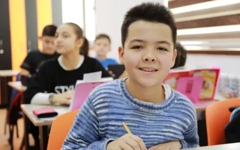 Uśmiechnięty chłopiec w swetrze, piszący w klasie. To ważny etap rozwoju 6 latka, nauka i nowe doświadczenia.