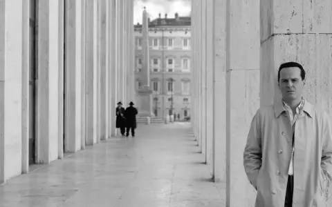 Un uomo in trench osserva l'obiettivo, mentre sullo sfondo due figure in abito scuro passeggiano sotto un colonnato. Potrebbe essere una scena da "Il talento di Mr. Ripley dove è stato girato".
