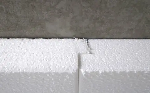 Styroporplatten werden zugeschnitten, um ein Kellerfenster zu isolieren. Die weißen Platten sind aneinandergefügt und grenzen an eine graue Betonfläche.