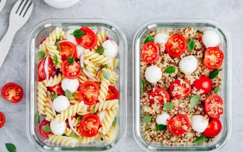 Zwei Glasbehälter mit frischen Mahlzeiten: links Nudelsalat, rechts ein bunter Quinoa-Salat mit Tomaten und Mozzarella.