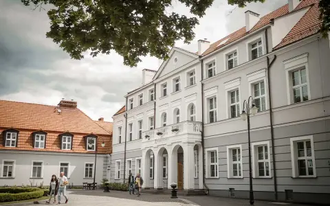 Elegancki pałac Zwartowo z białymi zdobieniami i łukowym wejściem. Na pierwszym planie para spaceruje po brukowanym dziedzińcu.