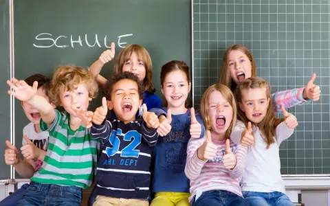 Fröhliche Kinder mit Daumen hoch vor einer Tafel mit dem Wort "SCHULE". Ein perfekter erster schultag!