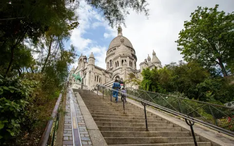 Dwie osoby wspinają się po schodach na wzgórze Montmartre, w tle Bazylika Sacré-Cœur.