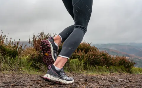 Gambe di una donna che fa trail run su un sentiero fangoso, con erica e colline nebbiose sullo sfondo.