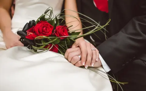 Dłonie nowożeńców splecione na tle bukietu czerwonych róż. Idealny moment, by pomyśleć, jak przygotować życzenia ślubne które zapadną w pamięć i wzruszą.