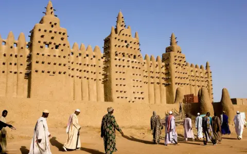 La grande mosquée de Djenné, chef-d'œuvre d'architecture en terre, se dresse sous un ciel bleu. Des hommes en tenues traditionnelles s'approchent, ajoutant vie à ce site historique.