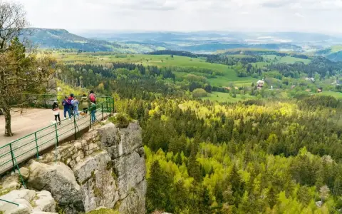 Grupa turystów podziwia rozległą panoramę Kotliny Kłodzkiej z platformy widokowej na skalistym zboczu.