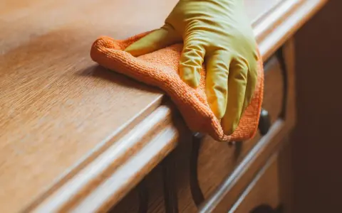 Main d'une personne portant un gant jaune, nettoyant un meuble en bois ciré encrassé avec un chiffon orange.