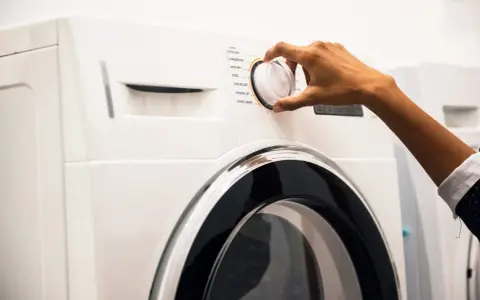 Main d'une personne ajustant le sélecteur d'un sèche-linge, choisissant un programme comme "Coton" ou "Laine". Les dimensions de la machine à laver sont standard.