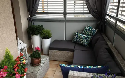 Przytulna zabudowa balkonu w bloku: rattanowy narożnik, poduszki w geometryczne wzory, rośliny i ozdobna latarenka tworzą relaksującą przestrzeń.