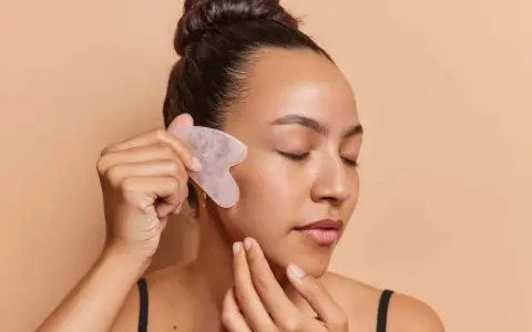 Frau mit geschlossenen Augen nutzt ein Gua Sha Stein zur Gesichtsmassage. Ob morgens oder abends, die Anwendung belebt die Haut.