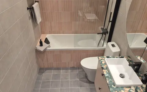 Ett modernt badrum med rosa kakel vid badkaret och grått klinkergolv. En vit toalett och ett handfat med en unik bänkskiva.