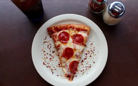 Kawałek pizzy pepperoni na talerzu, posypany płatkami chili i parmezanem. Idealny na szybki lunch, choć warto pamiętać o jego kcal.
