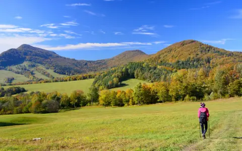 Jesień na **Beskid Niski szlaki**. Turystka w różowej kurtce idzie przez zieloną łąkę w kierunku kolorowych wzgórz.