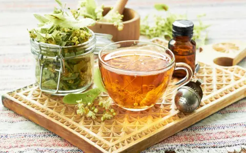 Tee aus Lindenblüten: welcher tee bei heiserkeit hilft, ist hier als dampfendes Getränk in einer Tasse, daneben getrocknete Blüten und ein Tropfenfläschchen.