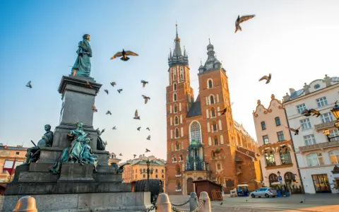 A monument and St. Mary's Basilica dominate this Krakow travel scene. Birds fly around the historic architecture under a clear sky.