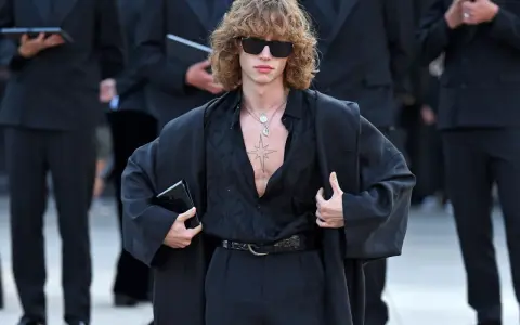 Ein Mann mit lockigem Haar und Sonnenbrille trägt eine schwarze, offen geknöpfte Bluse, weite Hosen und einen Mantel. Die Kleidung wirkt typisch 80er Jahre.