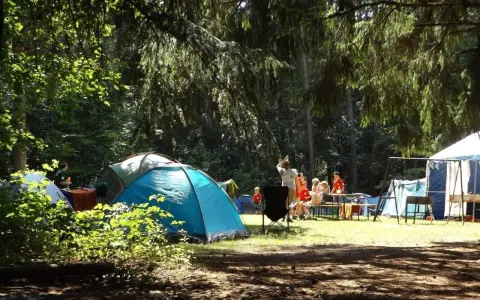 Słoneczny dzień na kempingu. Ludzie siedzą przy stole, rozmawiają, a obok rozstawione są namioty.