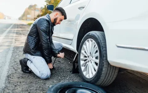 Mężczyzna klęczy przy białym samochodzie, używając podnośnika, by podnieść auto bez kanału. Obok leży zapasowa opona.