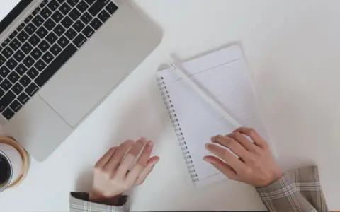 Ręce piszą CV po niemiecku przy laptopie, notatniku i kawie.