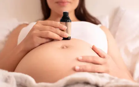 Schwangerenbauch mit einer Hand, die eine kleine Flasche Öl hält. Ab wann Dammmassage?