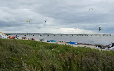 Kitesurfare förbereder sig vid Habo Ljung Camping. Många kites svävar i luften över en strand med en lång pir i bakgrunden.