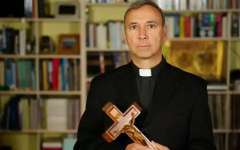 Duchowny z krzyżem w dłoniach, na tle regałów z książkami. Czy religia wlicza się do czerwonego paska?