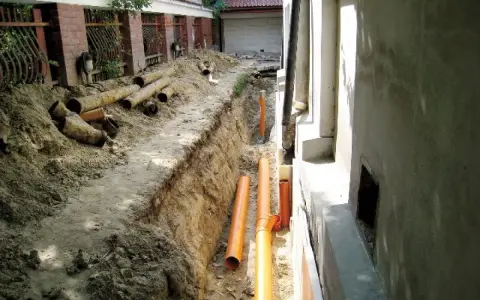 Trwa budowa drenażu wokół starego domu. W wykopie leżą nowe pomarańczowe rury i stare, zniszczone rury kanalizacyjne.