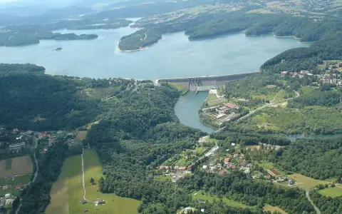 Widok z lotu ptaka na zapora wodna w Myczkowcach, otoczona zielonymi wzgórzami i rozległym jeziorem.