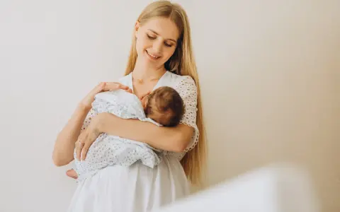 Uśmiechnięta młoda mama tuli noworodka, karmiąc go piersią. Delikatne spojrzenie i biała sukienka podkreślają jej macierzyństwo.