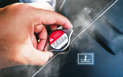 Dłoń dotyka gorącego korka chłodnicy, z którego unosi się para. Widoczny symbol ostrzegawczy i napis "DANGER WARNING". Kuhlmittel stand jest wysoki.