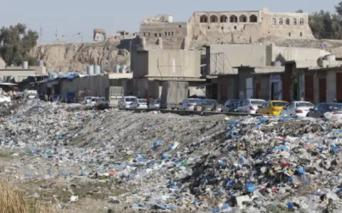 Müllberge säumen die Straßen von Kirkuk, Irak. Autos parken hinter den Geschäften, während im Hintergrund alte Gebäude auf einem Hügel stehen.