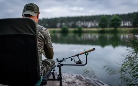 Wędkarz w kamuflażu siedzi na krześle nad jeziorem, gotowy do łowienia. Jego wędka feeder 3m spoczywa na podpórce, czekając na branie.