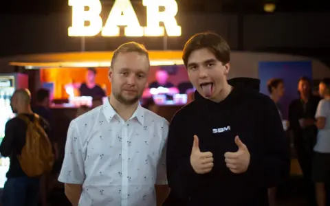 Dwóch mężczyzn pozuje przed napisem "BAR". Jeden w koszuli w palmy, drugi w bluzie z napisem SBM, pokazuje kciuki w górę i wystawia język.