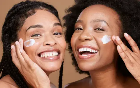 Deux femmes souriantes appliquent une crème sur leur visage, un geste pour une exfoliation visage réussie.