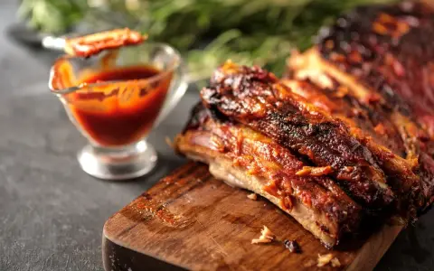 Costine al forno succulente e caramellate, servite su tagliere di legno con salsa barbecue in ciotolina.