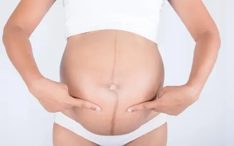 Ciemna linia, linea negra, biegnie przez brzuch ciężarnej kobiety, która wskazuje na nią palcami.