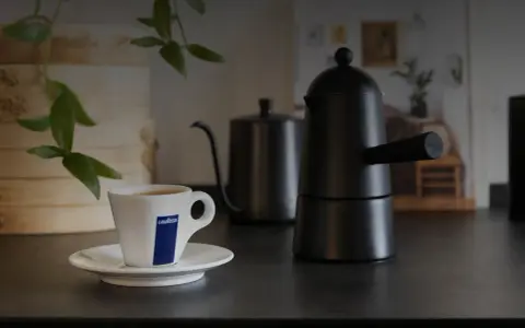 Eine Tasse Lavazza-Kaffee steht neben einer schwarzen Mokkakanne. Die Szene vermittelt die klassische italienische Kaffeezubereitung.