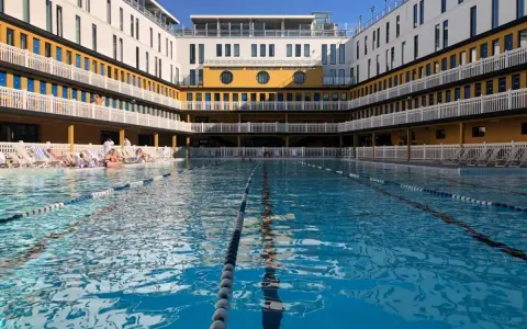 Piscine art déco parisienne, avec ses bâtiments jaunes et blancs, ses balcons et ses transats. L'eau bleue invite à la baignade.