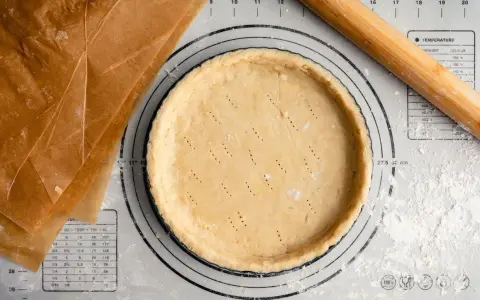 Pâte sucrée noisette prête à être garnie, sur un tapis de pâtisserie avec un rouleau à bois et du papier sulfurisé.