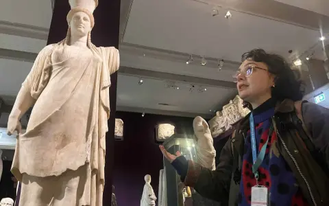 A woman with a museum pass for Turkey gestures towards a marble statue of a woman in a museum.