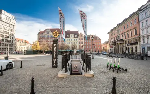 Wejście do podziemi z napisem "Movie Gate" i dwoma pionowymi banerami. W tle kolorowe kamienice i błękitne niebo.