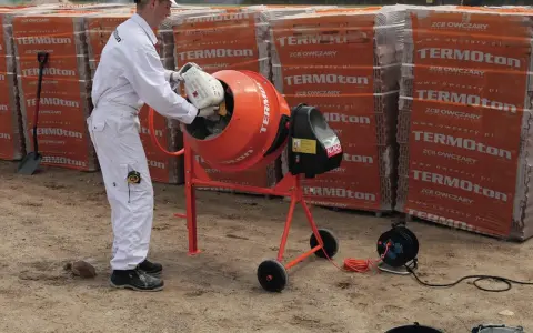 Mężczyzna w białym kombinezonie wsypuje składniki do betoniarki, przygotowując beton B30. W tle palety pustaków.