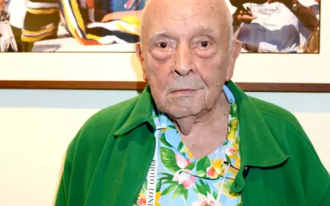 An elderly man with a green jacket over a floral shirt. This image might spark some david bailey interesting facts about his iconic style.