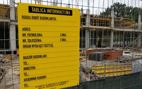 Żółta tablica informacyjna budowy z danymi o pozwoleniu, adresie i kontaktach. W tle widać konstrukcję budynku.