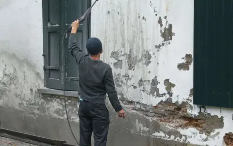 Un homme utilise un nettoyeur haute pression pour le sablage facade, révélant la brique sous l'enduit décrépit.