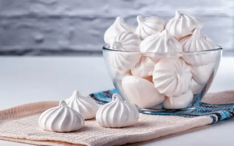 Douceur aérienne : des meringues délicates, comme préparées au bain-marie, dans un bol en verre et sur un tissu texturé.