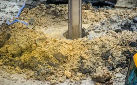 Wiertnica zagłębia się w ziemię, tworząc otwór pod fundamenty palowe. Wokół rozsypana glina i kamienie.