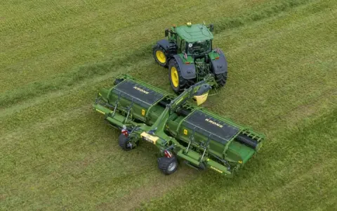 Zielony traktor z podłączoną zgrabiarką taśmową Krone pracuje na zielonym polu, zbierając skoszoną trawę.